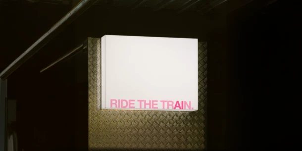 Image of a rectangular lit sign set on a sheet of corrugated metal. The light is bright white and the text is in all caps in pink with the letters "AI" subtly set apart in a darker pink.. TEXT: "RIDE THE TRAIN."